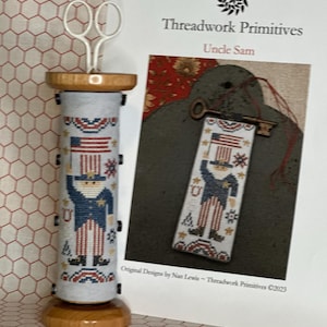 May include: A cylindrical wooden spool stand holding a cross-stitch fabric piece featuring an Uncle Sam design with an American flag, stars, and red, white, and blue motifs. White scissors sit on top. Beside it is a pattern sheet titled Threadwork Primitives.