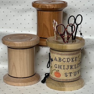 May include: Three wooden spool-shaped containers, two are natural wood with black trim, one is natural wood with a black trim and a band of cross-stitch fabric with the alphabet and a pumpkin design. The container with the fabric band has two pairs of scissors and a small earring inside.
