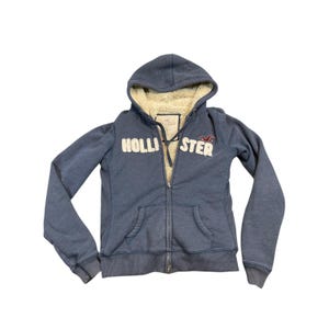 May include: A blue zip-up hoodie featuring a cream-colored sherpa lining inside the hood and body. The front displays the white embroidered text HOLLISTER with a small red bird logo above the right side of the text and two front pockets.