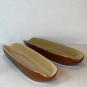May include: Two brown and beige ceramic serving dishes shaped like corn on the cob. The dishes have a textured surface and are perfect for serving hot dogs or other finger foods.