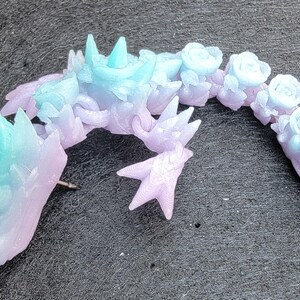 Pu&ograve; includere: Una scultura in resina di un drago di colore blu pastello e viola con dettagli floreali. Il drago &egrave; arrotolato su se stesso con la testa appoggiata sulla coda.