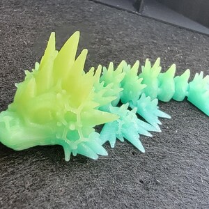 Pu&ograve; includere: Una creatura stampata in 3D con un corpo spinoso e cristallino. La creatura &egrave; verde e blu, con un effetto sfumato dal verde al blu. La creatura &egrave; sdraiata su una superficie grigio scuro.