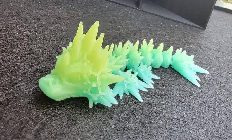 Pu&ograve; includere: Una creatura stampata in 3D con un corpo spinoso e cristallino. La creatura &egrave; verde e blu, con un effetto sfumato dal verde al blu. La creatura &egrave; sdraiata su una superficie grigio scuro.