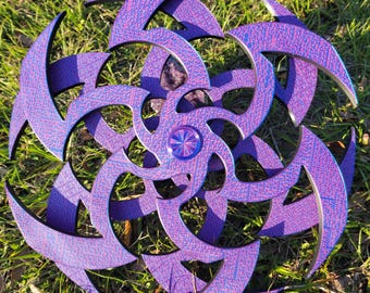 May include: A purple and blue iridescent wooden starburst with a central metal disc. The starburst is made of multiple layers of wood, each with a different pattern.