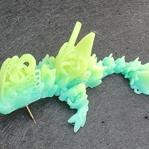 Pu&ograve; includere: Una statuetta di drago stampata in 3D di colore verde e blu con dettagli intricati. Il drago &egrave; sdraiato sul fianco con la testa girata a sinistra. La statuetta &egrave; realizzata in un materiale traslucido e presenta un effetto sfumato di colore.