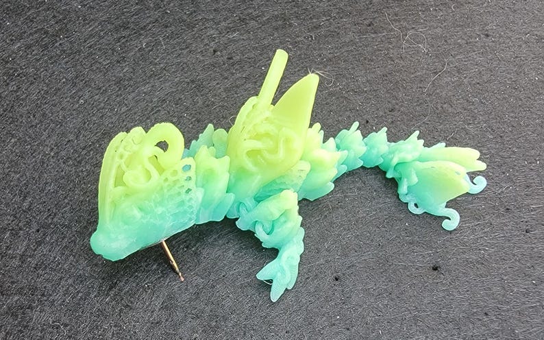 Pu&ograve; includere: Una statuetta di drago stampata in 3D di colore verde e blu con dettagli intricati. Il drago &egrave; sdraiato sul fianco con la testa girata a sinistra. La statuetta &egrave; realizzata in un materiale traslucido e presenta un effetto sfumato di colore.