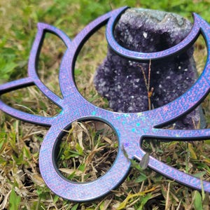 May include: A decorative wand with a floral design. The wand is a gradient of purple and blue with a glittery finish and black trim. The wand is lying on a bed of grass, with a purple crystal in the background.