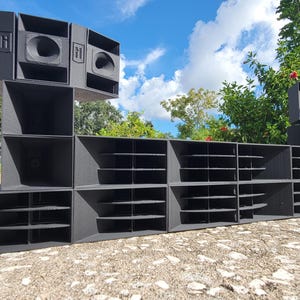 May include: A large, modular black speaker system with multiple components. The system features various speaker enclosures, including horn-loaded and vented designs. The speakers are arranged in a stacked and horizontal configuration, set against a backdrop of a blue sky and greenery.