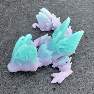 Pu&ograve; includere: Una piccola statuetta di una creatura alata, stampata in 3D, con dettagli intricati. La statuetta &egrave; realizzata in plastica trasparente con un gradiente di colori che va dal viola al turchese.