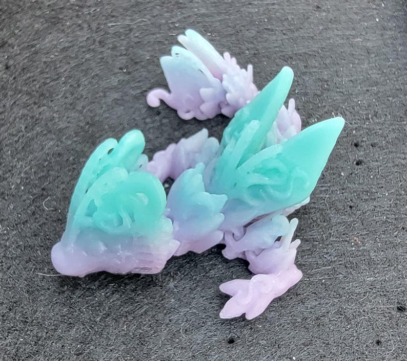 Pu&ograve; includere: Una piccola statuetta di una creatura alata, stampata in 3D, con dettagli intricati. La statuetta &egrave; realizzata in plastica trasparente con un gradiente di colori che va dal viola al turchese.