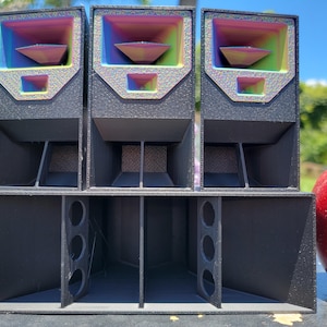 May include: Three black speaker enclosures with rainbow-colored interior accents. The speakers are arranged in a stacked configuration, with a large red apple visible on the right side of the image. The speakers have a modern, geometric design.