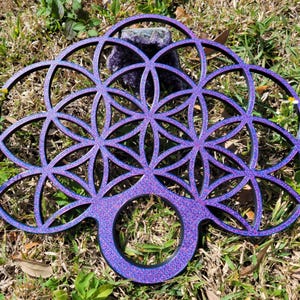 May include: A purple and blue decorative wooden wall hanging featuring the Flower of Life geometric pattern. The design has a circular handle at the bottom and is placed on a bed of grass.