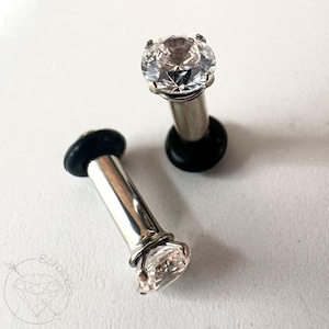 May include: Two silver-colored ear gauges with clear, faceted crystal tops. The gauges have a cylindrical post and a black rubber O-ring on the back. One gauge is angled, and the other is upright. The jewelry is on a white background.