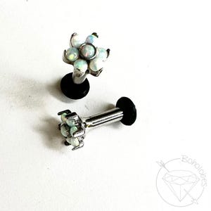 May include: Two silver-colored lip piercing studs with a flower design. Each flower is made of multiple white opal-like stones. The studs have a black backing. The image is on a white background.