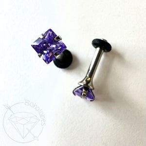May include: Two pieces of body jewelry. One is a black plug with a square, faceted, purple gemstone. The other is a silver-colored barbell with a purple gemstone at the end. The jewelry is on a white background.