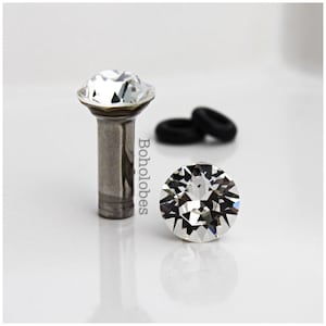 May include: A metal stud earring with a clear, faceted, round crystal top and a cylindrical post. The word Boholobes is printed vertically next to the stud. Two small black rubber earring backs are positioned in the background behind the stud.