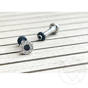 May include: Two silver-colored barbell earrings with a black center and a ring of clear crystals. The earrings have a cylindrical post and a round, flat end. One earring is in focus, the other is slightly blurred in the background.