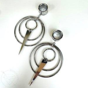 May include: A pair of silver-toned ear gauges with dangling geometric designs. Each gauge has a circular plug connected to a series of concentric rings and a triangular pendant. The jewelry is displayed on a white surface.