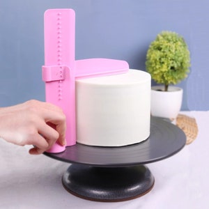 May include: A pink cake leveler is used to smooth the white frosting on a round cake. The cake sits on a dark gray cake stand. The leveler has a vertical ruler with measurement markings.