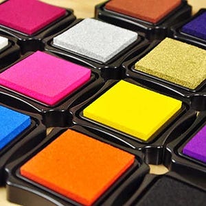 Könnte beinhalten: Eine Sammlung farbenfroher Stempelkissen in einer Vielzahl von leuchtenden Farben, darunter Pink, Gelb, Orange und Blau. Jedes Kissen ist quadratisch und in einer schwarzen Kunststoffschale angeordnet. Die Stempelkissen sind für Bastel- und Kunstprojekte konzipiert.