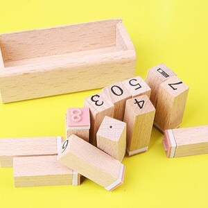 May include: A set of wooden number stamps in a wooden box. The stamps are arranged in a pile, with the numbers 1, 2, 3, 4, 5, 6, 7, 8, and 9 visible. The stamps are all made of natural wood and have a light brown color.