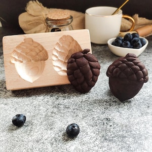 May include: A wooden mold with two acorn shapes, one filled with a dark brown chocolate acorn and the other with a dark brown chocolate pine cone. Two blueberries are in the foreground.