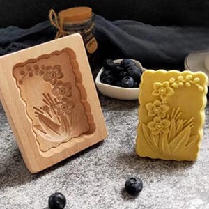 May include: A wooden mold for making soap or cake, with a floral design. The mold is next to a yellow soap bar with the same floral design.