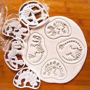 Könnte beinhalten: Ein Set mit sechs weißen Plastik-Ausstechern in Form von Dinosauriern. Die Ausstecher sind in Form verschiedener Dinosaurier, darunter ein T-Rex, ein Stegosaurus und ein Triceratops. Die Ausstecher befinden sich auf einer weißen Oberfläche mit einem hellbraunen Hintergrund.