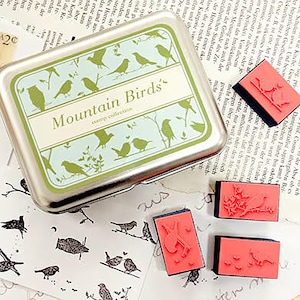 Könnte beinhalten: Eine silberne Metallbox mit einem grünen und blauen Design mit dem Text "Mountain Birds' stamp collection". Die Box enthält vier rote Gummistempel mit verschiedenen Vogel- und Blumenmotiven.