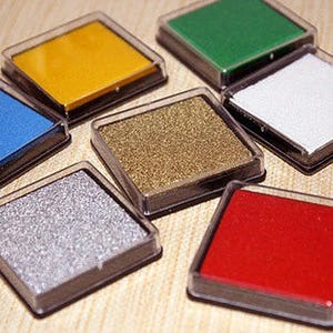 Könnte beinhalten: Sortiment von sechs Stempelkissen in verschiedenen Farben, darunter Gold, Silber, Rot, Grün, Blau und Gelb. Die Stempelkissen befinden sich in durchsichtigen Plastikbehältern mit schwarzem Rand.