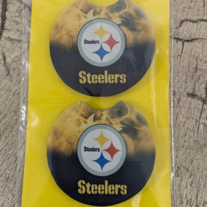 May include: Two black and yellow round drink coasters with a Steelers logo and the word "Steelers" printed on them. The coasters have a yellow and black flame design.