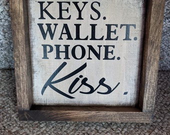May include: A wooden framed sign with the text "KEYS. WALLET. PHONE. Kiss." in black lettering on a white background.
