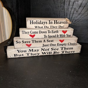 May include: A set of four wooden signs with a whitewashed finish and black lettering. The signs are stacked on top of each other and feature a message about holidays in heaven. The signs are decorated with red hearts.