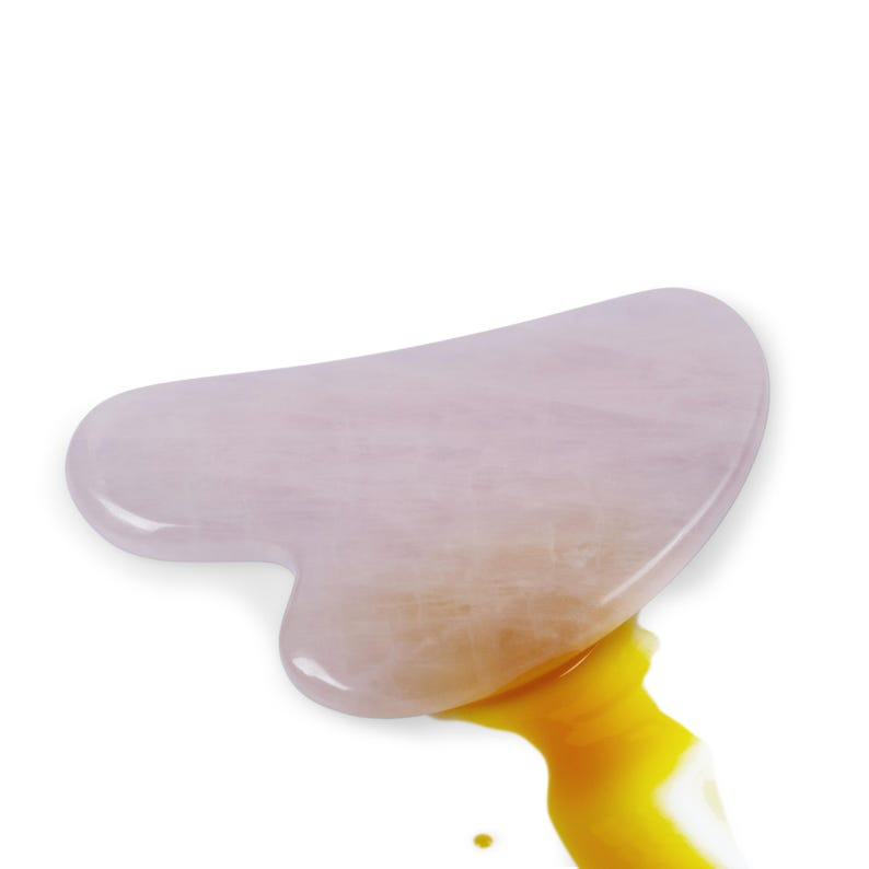 May include: A smooth, heart-shaped gua sha tool crafted from pale pink rose quartz stone. The surface of the stone displays a natural, semi-translucent crystalline texture. A small amount of golden yellow oil is visible beneath the bottom edge.