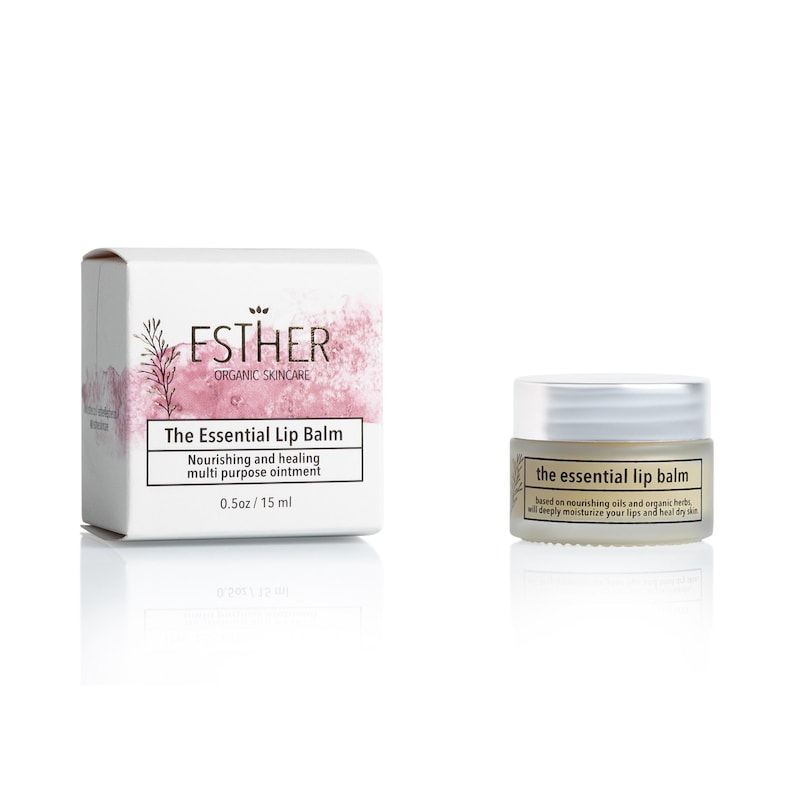 Puede incluir: Una caja de cart&oacute;n blanca y un peque&ntilde;o frasco de vidrio de b&aacute;lsamo labial Esther Organic Skincare The Essential Lip Balm. La caja tiene detalles en acuarela rosa y texto que indica 15 ml. El frasco tiene tapa plateada y etiqueta negra.