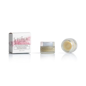 Puede incluir: Una caja de cart&oacute;n blanca con detalles florales rosas, un frasco peque&ntilde;o con tapa plateada y un frasco abierto con b&aacute;lsamo labial amarillo p&aacute;lido. Las etiquetas dicen Esther Organic Skincare The Essential Lip Balm 0.5oz / 15ml.