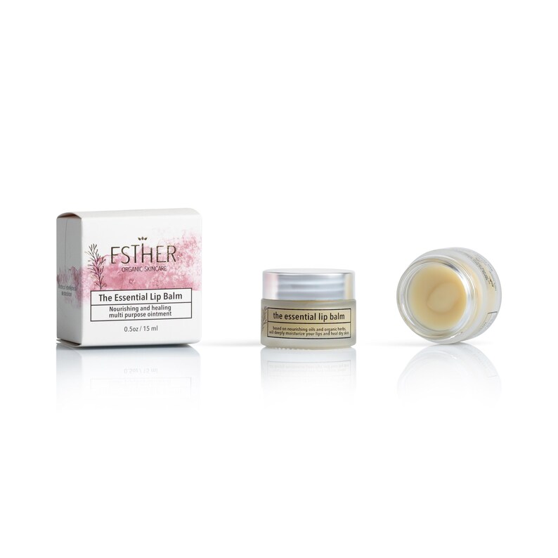 Puede incluir: Una caja de cart&oacute;n blanca con detalles florales rosas, un frasco peque&ntilde;o con tapa plateada y un frasco abierto con b&aacute;lsamo labial amarillo p&aacute;lido. Las etiquetas dicen Esther Organic Skincare The Essential Lip Balm 0.5oz / 15ml.