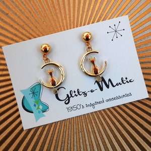 Könnte beinhalten: Goldfarbene Ohrringe mit einem Halbmond-Design und einer kleinen Katzensilhouette. Die Ohrringe befinden sich auf einer weißen Karte mit dem Text "Glitz-a-Matic 1950's inspired accessories".
