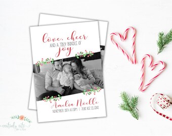 May include: A birth announcement card with a black and white family photo, red text, and floral accents. The card reads "love, cheer, and a tiny bundle of joy" and includes the baby's name, birth date, and weight. Candy canes and greenery are arranged around the card.