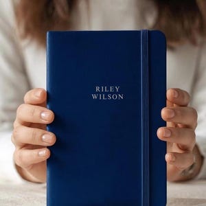 May include: A navy blue notebook with the text "RILEY WILSON" in white lettering. The notebook has a matching elastic band closure. The image shows the notebook being held by a person.