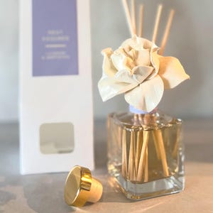 May include: A clear glass diffuser bottle with a white flower and wooden reeds. A white box with text and a gold-topped stopper are next to the bottle. The text on the box reads "REED DIFFUSER" and "LAVENDER & COTTON".