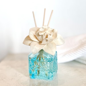 May include: A white ceramic flower diffuser with wooden sticks in a blue glass bottle with a diamond pattern. The diffuser is on a white surface.
