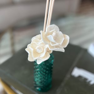May include: A teal glass reed diffuser bottle with a textured diamond pattern, containing two cream-colored wooden flowers and three thin, light-colored wooden reeds extending upward from the narrow neck of the glass vessel.