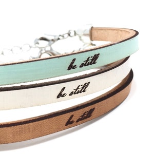 May include: Close-up of three leather bracelets in pastel colors: mint green, white, and tan. Each bracelet is engraved with the words "be still" in black script. The bracelets are connected by a silver chain.