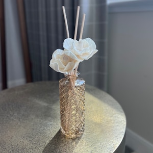 May include: A reed diffuser set featuring a cylindrical glass bottle with a textured diamond-patterned surface in a light amber hue. Three thin wooden reeds are inserted into the bottle, topped with two cream-colored, layered floral-shaped diffusers.