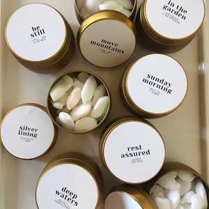 May include: A collection of gold metal tins with white labels and white wax melts inside. The labels have different names and scents, including "be still", "move mountains", "in the garden", "sunday morning", "silver lining", "rest assured", "deep waters", and "abiding".