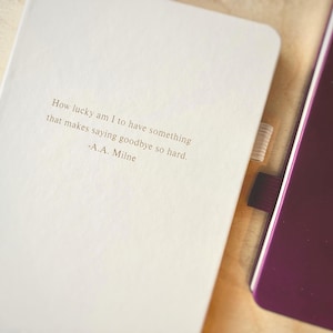 May include: Two notebooks, one white and one burgundy, are displayed. The white notebook features the quote "How lucky am I to have something that makes saying goodbye so hard." by A.A. Milne. The burgundy notebook is partially visible.