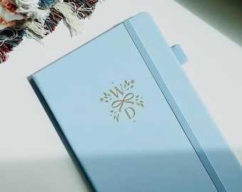 Puede incluir: Un cuaderno azul claro con una banda elástica de cierre vertical y un lazo para bolígrafo. La cubierta presenta un monograma en lámina dorada con las letras W y D rodeadas por un diseño botánico. Las palabras COMMON NOTES están impresas abajo.