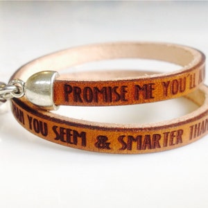 May include: A light brown leather wrap bracelet with the engraved text "PROMISE ME YOU'LL ALWAYS BE SMARTER THAN YOU SEEM". The bracelet has a silver-colored clasp and connector. The leather band is approximately 0.5 inches wide.