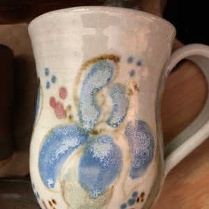 以下が含まれることがあります： 青と緑の花柄が描かれた白い陶器のマグカップ。マグカップは丸みを帯びたボディとハンドルが付いています。